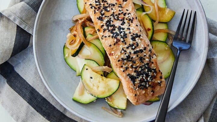 A plate with a cooked salmon fillet topped with black and white sesame seeds, served over sautéed zucchini and onions, makes a perfect March Mediterranean Diet Dinner—finished with a black fork on the side.