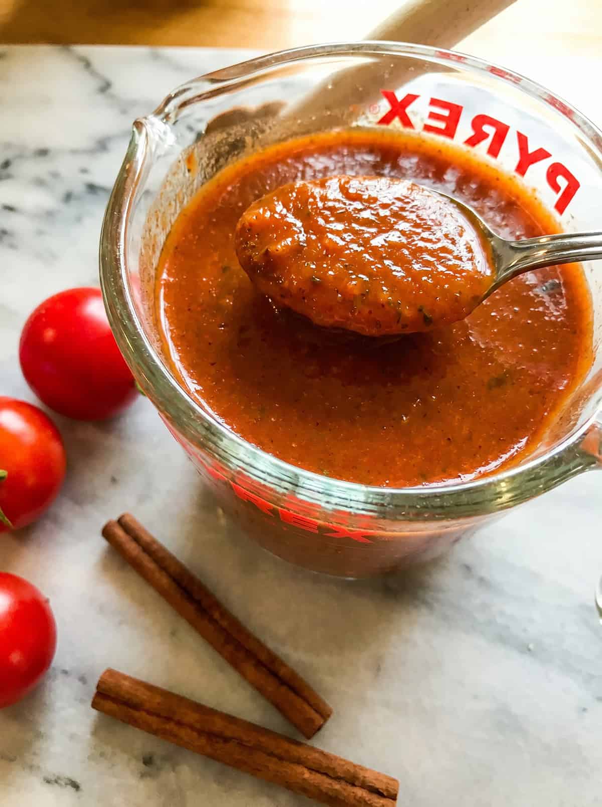 A glass measuring cup filled with enchilada sauce, a spoon resting on top, fresh tomatoes, and two cinnamon sticks on a marble surface.