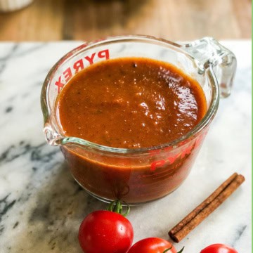 A glass measuring cup filled with thick brown-red enchilada sauce sits on a marble surface next to fresh tomatoes and a cinnamon stick.
