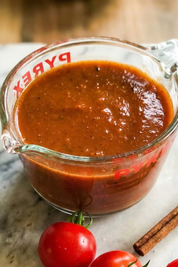 A glass measuring cup filled with thick brown-red enchilada sauce sits on a marble surface next to fresh tomatoes and a cinnamon stick.