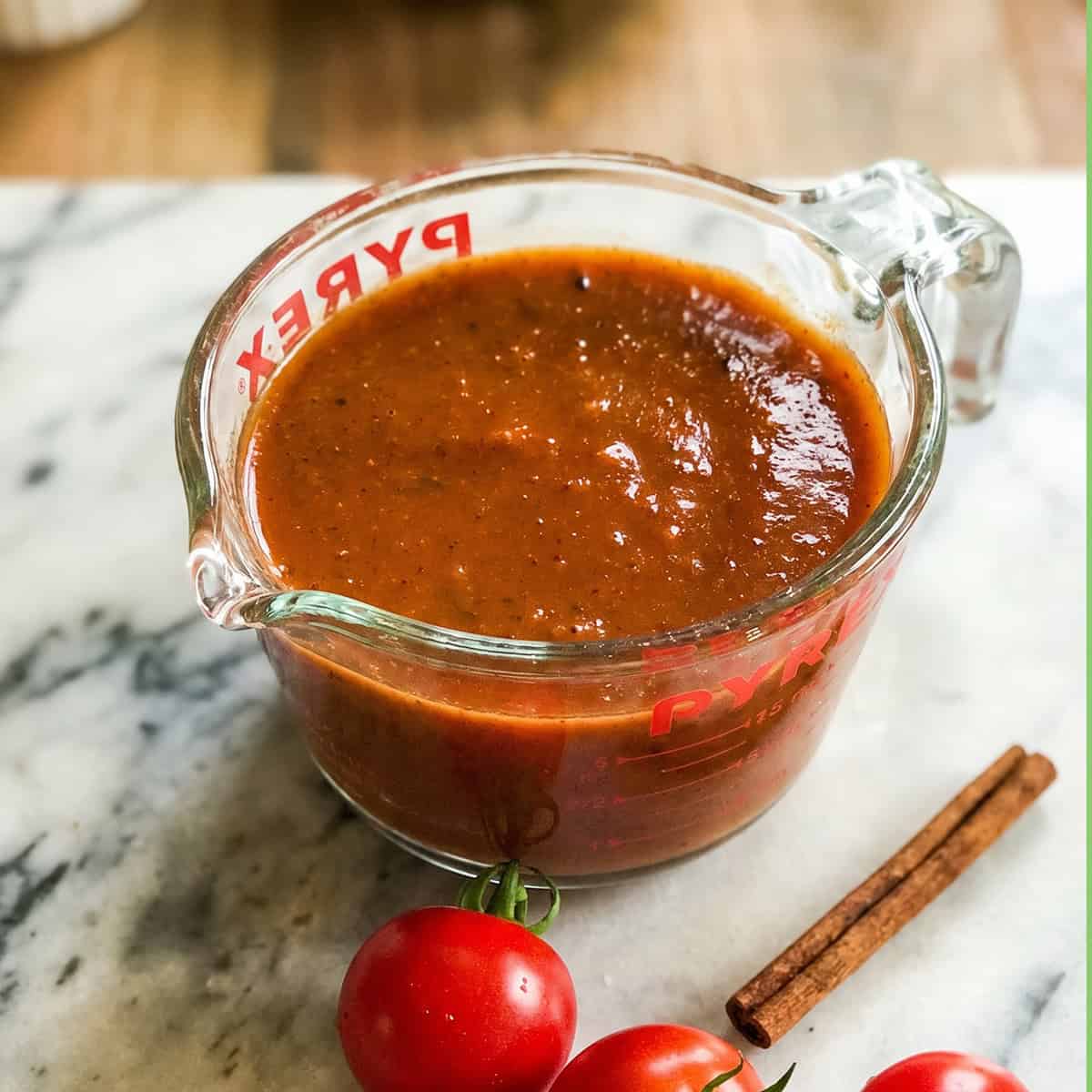 A glass measuring cup filled with thick brown-red enchilada sauce sits on a marble surface next to fresh tomatoes and a cinnamon stick.