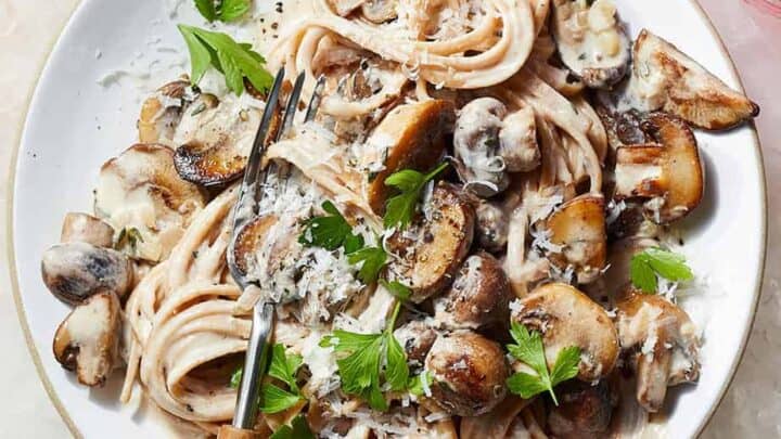A plate of creamy pasta topped with sautéed mushrooms, grated cheese, and fresh parsley makes for one of those comforting high protein dinners, with a fork resting on the side.