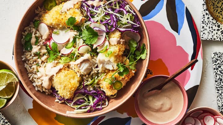 A high protein dinner featuring a bowl of rice topped with breaded chicken, shredded cabbage, radishes, cilantro, and creamy sauce, served on a colorful floral plate with sides of lime, radishes, and extra sauce.
