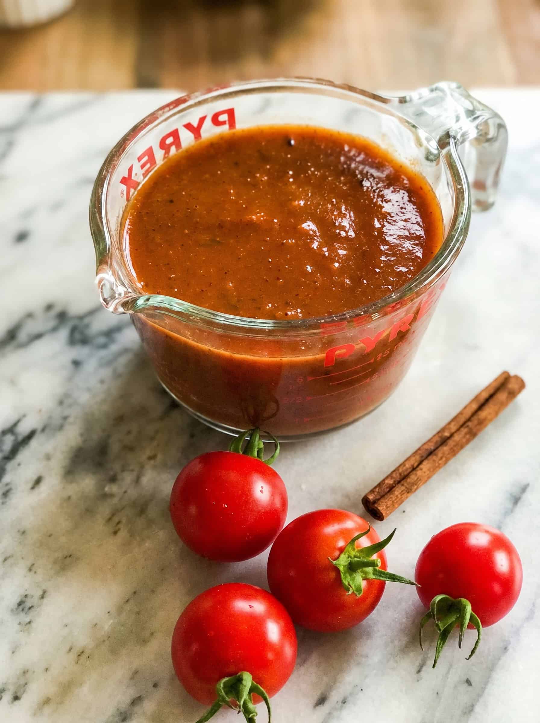 A glass measuring cup filled with enchilada sauce sits on a marble surface next to three fresh tomatoes and two cinnamon sticks.