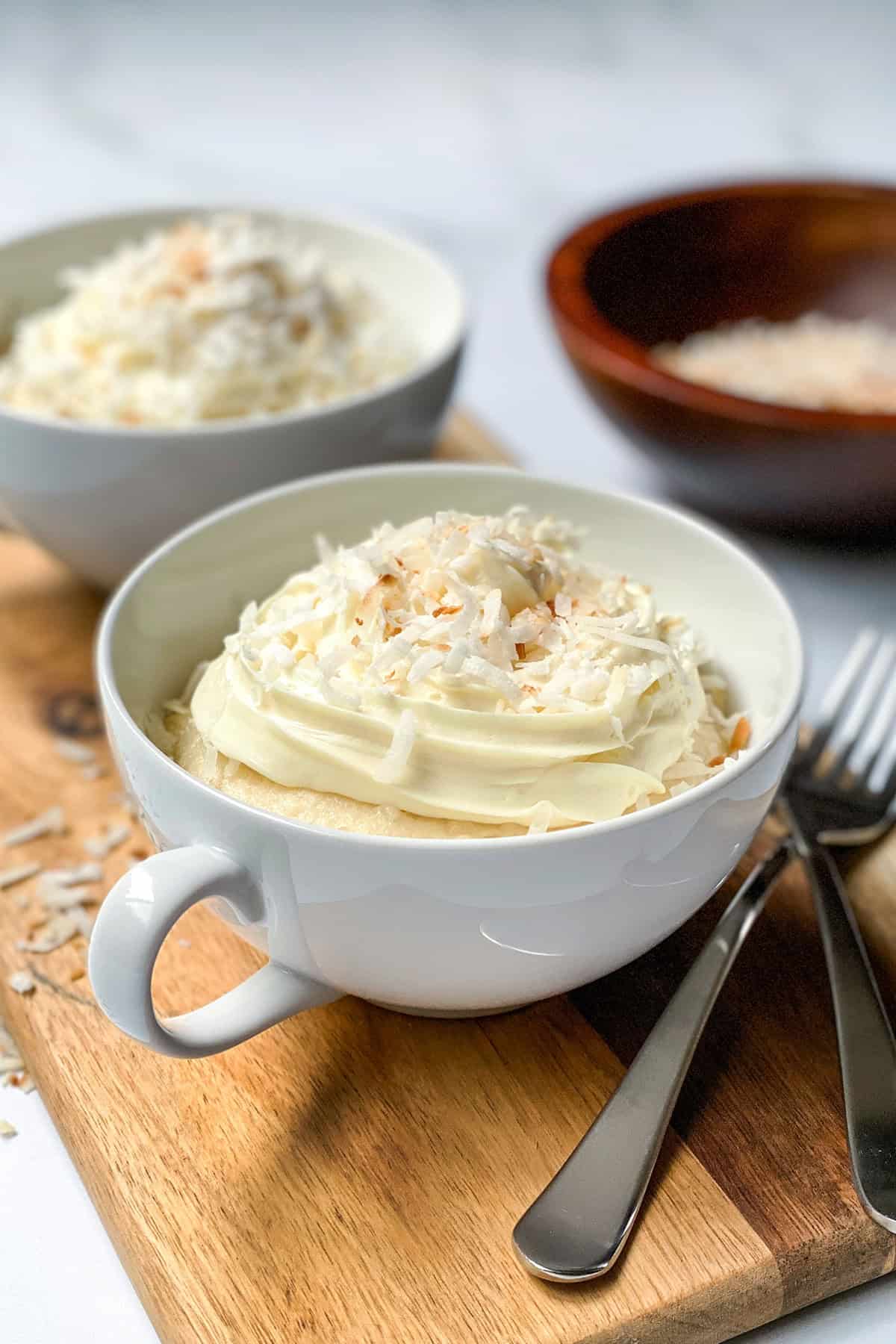 A coconut mug cake in a white cup, topped with white frosting and shredded coconut, sits on a wooden board, with bowls of shredded coconut and two forks beside it.