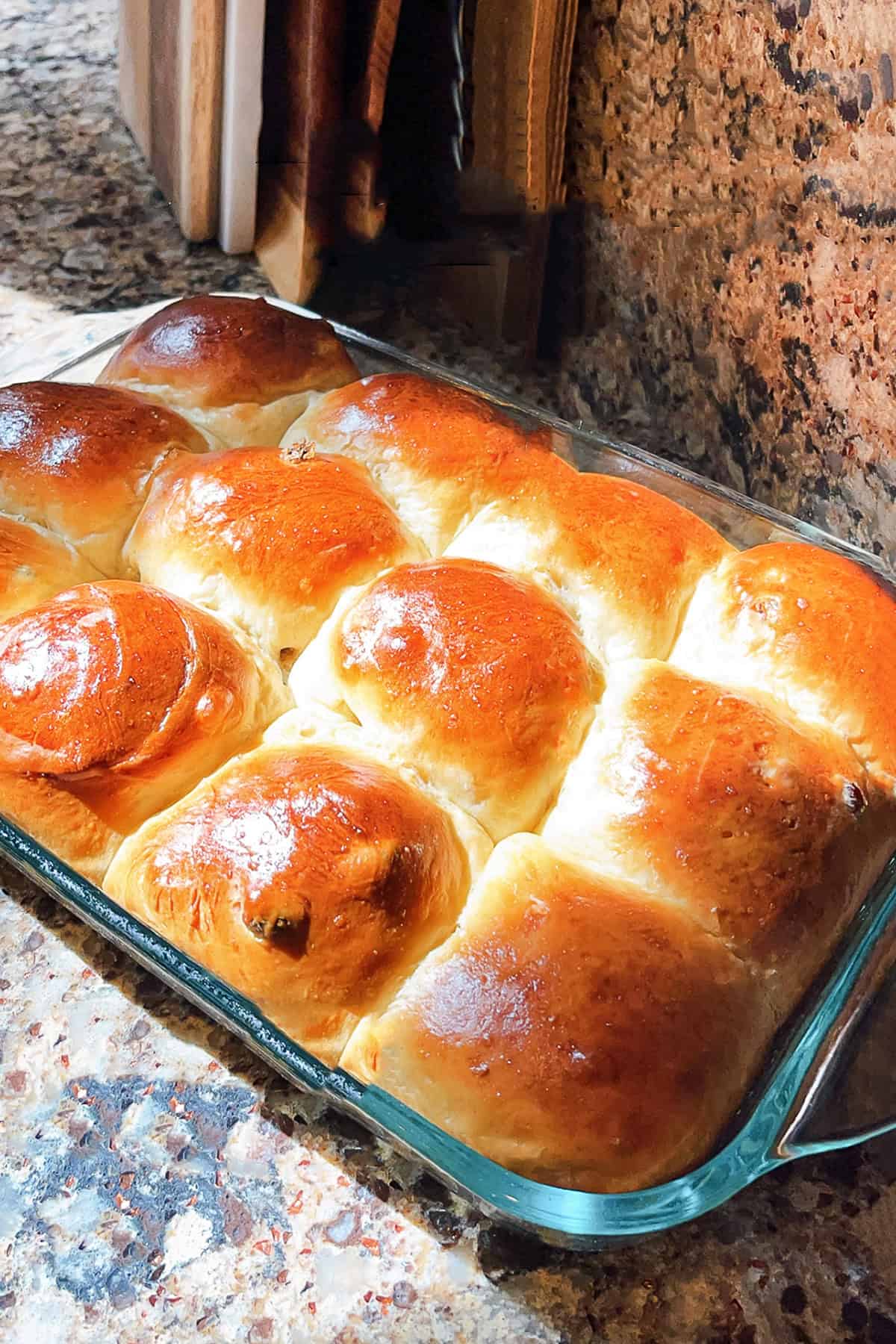 A glass baking dish filled with nine golden-brown hot cross buns sits on a speckled granite countertop by a backsplash and wooden utensils.