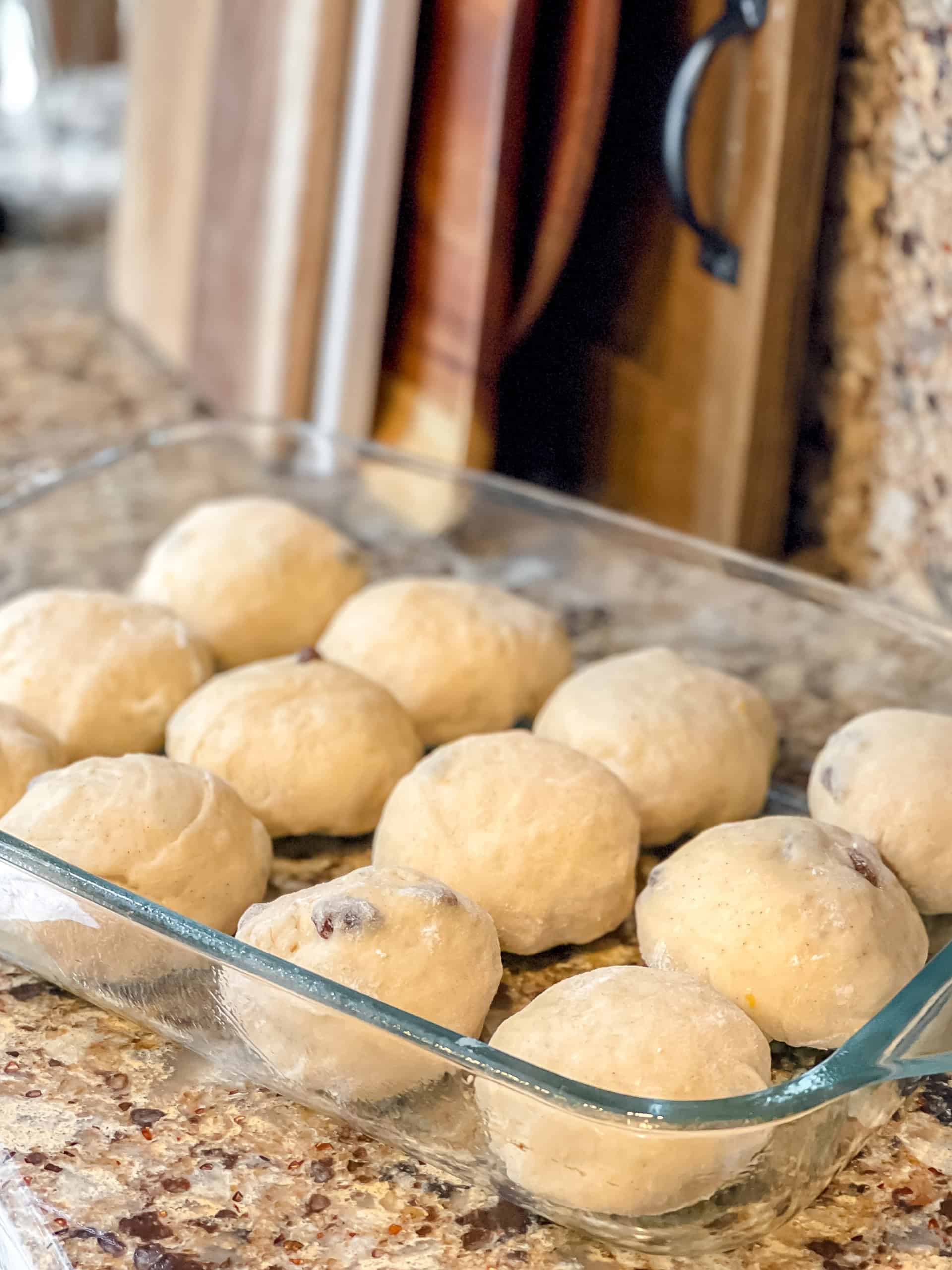 A glass baking dish with twelve unbaked dough balls for a hot cross buns recipe rests on a granite countertop near wooden cutting boards.