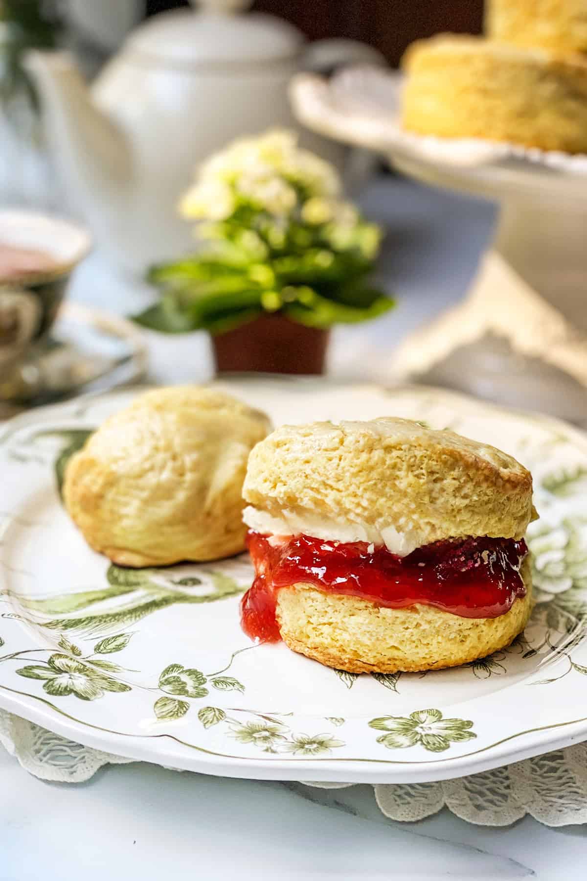 A scone split in half with cream and strawberry jam, placed on a floral plate beside a plain scone; a teapot and flowers are in the background, evoking the charm of an authentic Irish scones recipe.