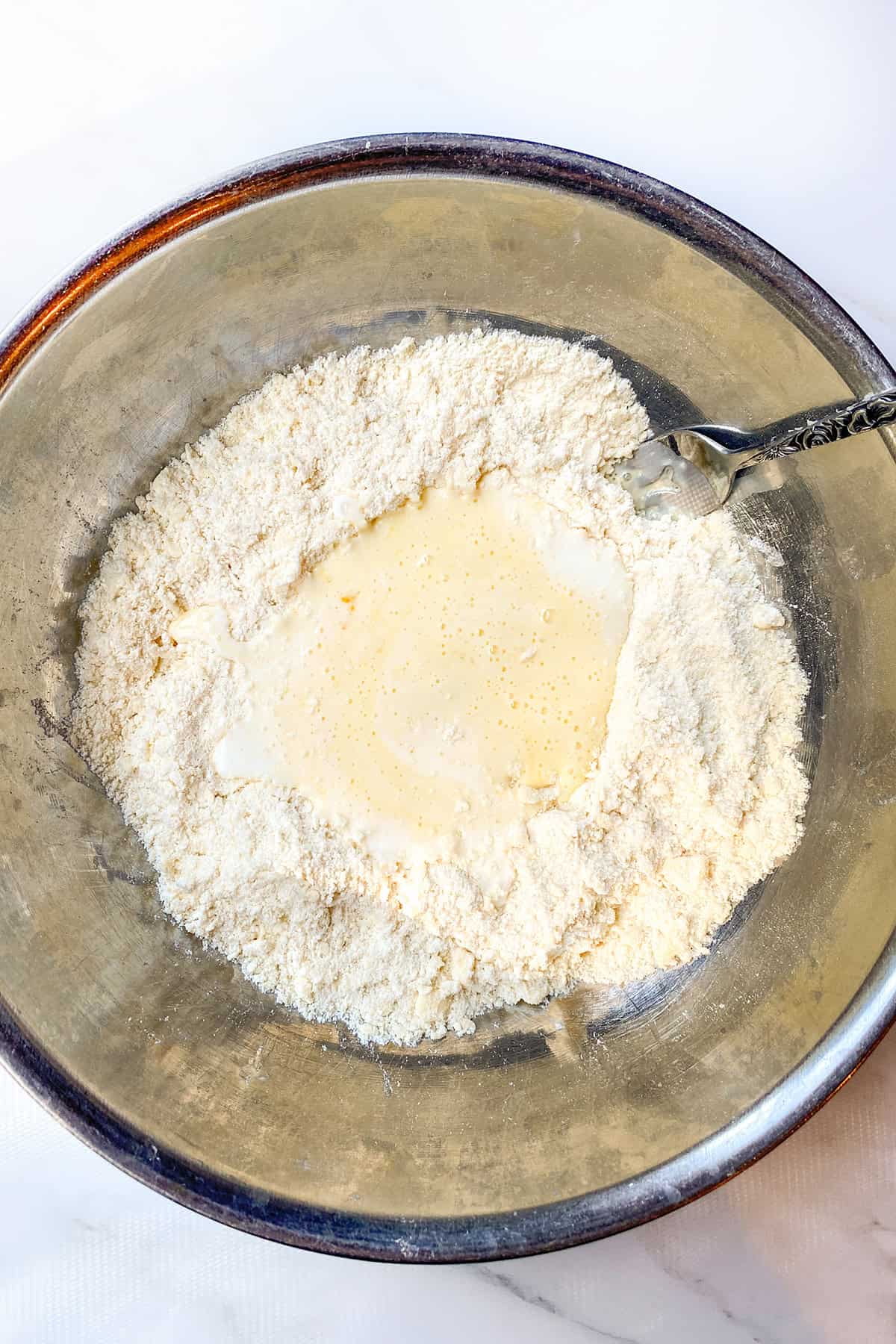 A metal bowl contains flour with milk and eggs in the center, partially mixed with a fork-just the beginning steps of an Irish scones recipe.