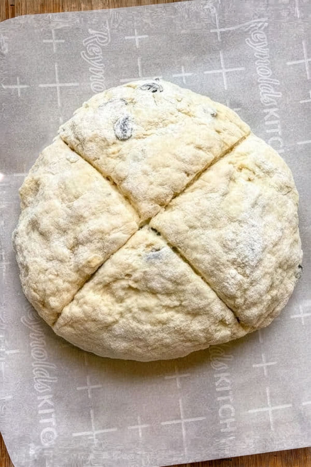 Round, unbaked Irish Soda Bread dough with a cross shape cut into the top, resting on parchment paper with a faint grid pattern.