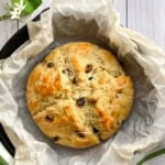 A round loaf of Irish Soda Bread, studded with raisins, sits on parchment paper in a baking pan, boasting a golden crust and a beautifully scored top.