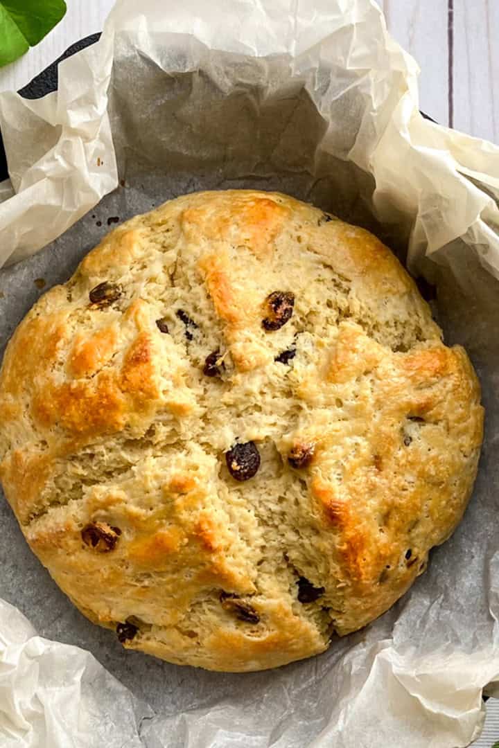 A round loaf of Irish Soda Bread, studded with raisins, sits on parchment paper in a baking pan, boasting a golden crust and a beautifully scored top.