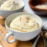 A white cup filled with coconut mug cake, topped with whipped cream and shredded coconut, sits on a wooden board with forks and a bowl of coconut in the background.