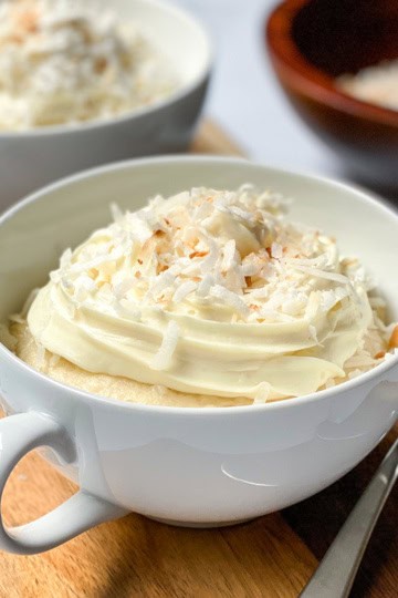 A white cup filled with coconut mug cake, topped with whipped cream and shredded coconut, sits on a wooden board with forks and a bowl of coconut in the background.