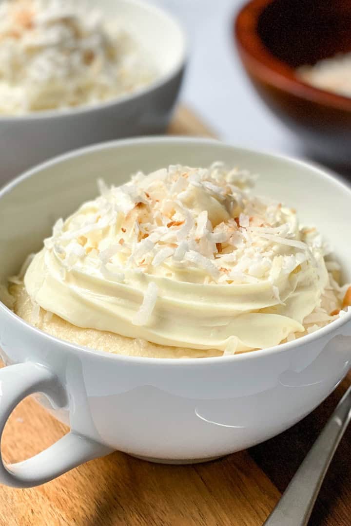 A white cup filled with coconut mug cake, topped with whipped cream and shredded coconut, sits on a wooden board with forks and a bowl of coconut in the background.