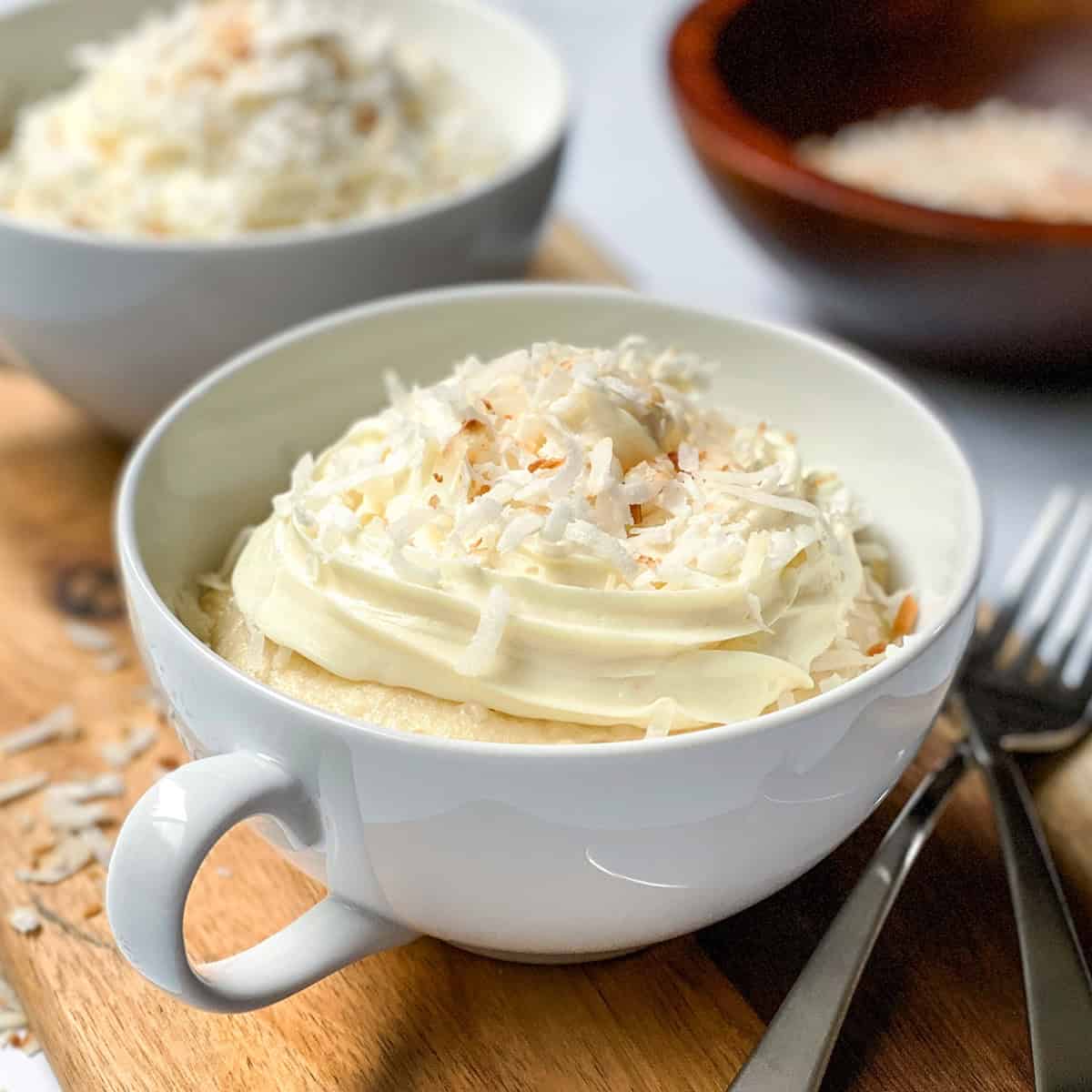 A white cup filled with coconut mug cake, topped with whipped cream and shredded coconut, sits on a wooden board with forks and a bowl of coconut in the background.