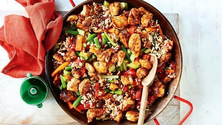 A skillet filled with cooked rice, chicken pieces, and mixed vegetables makes one of the perfect 5 Ingredient Spring Dinners, with a wooden spoon resting on the side and a red cloth nearby.