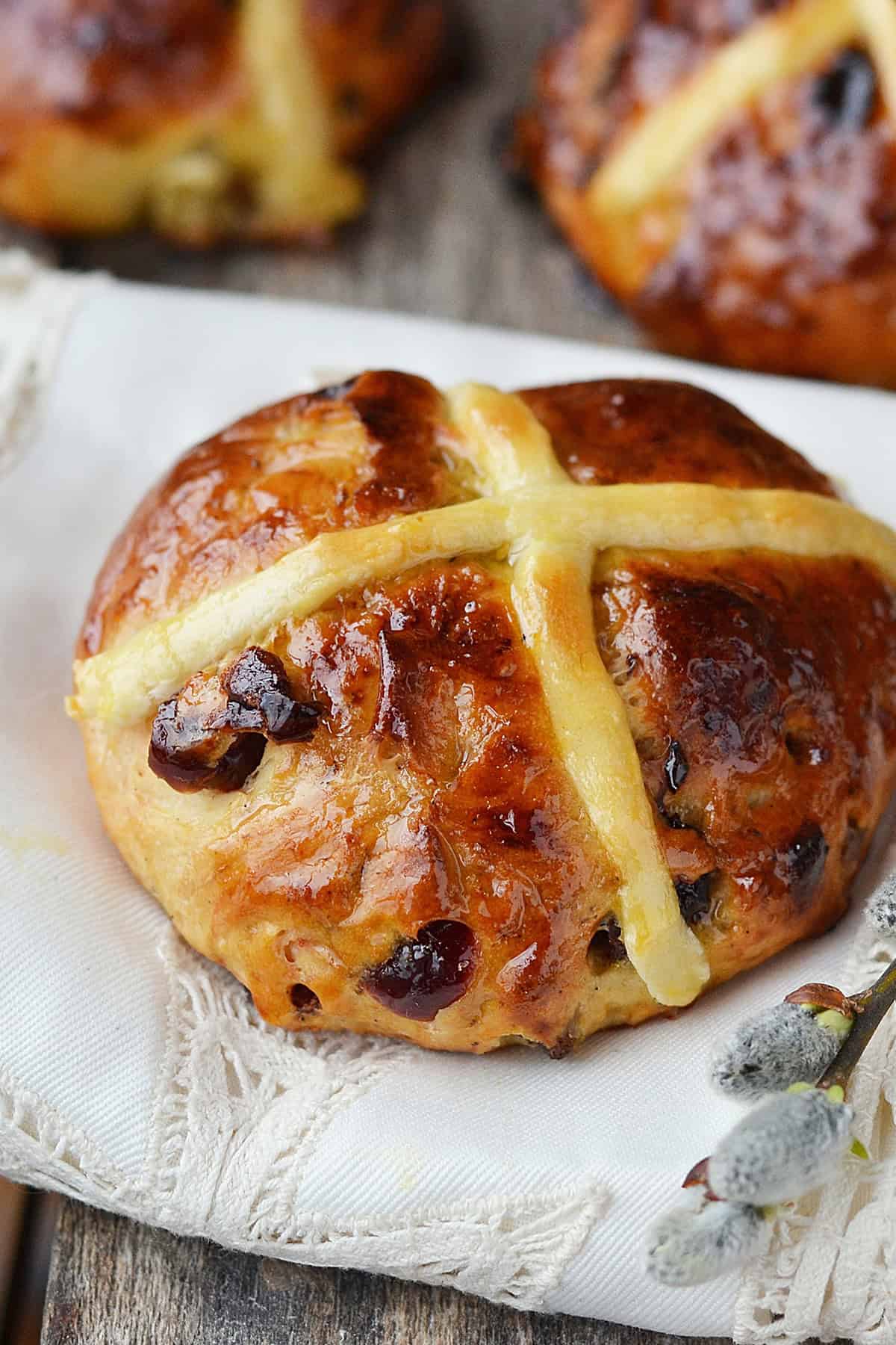 A close-up of a glazed hot cross bun, made from a traditional hot cross buns recipe, with a yellow cross on top, resting on a white cloth and dotted with visible raisins or dried fruit pieces.