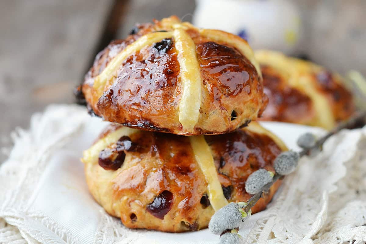 Two glazed hot cross buns with raisins, stacked on a white cloth, with a sprig of pussy willow beside them-a perfect scene to inspire your next hot cross buns recipe.