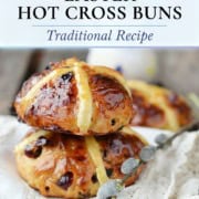 Three shiny, golden-brown hot cross buns with icing crosses are stacked on a cloth, with a decorative sprig beside them. Text above promotes a classic hot cross buns recipe for your traditional Easter celebration.
