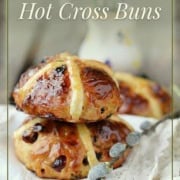 Two glazed hot cross buns with icing crosses are stacked on a cloth, accompanied by pussy willow branches. Text above reads "MAKE AHEAD Hot Cross Buns." Try this classic hot cross buns recipe for a delicious treat any time.