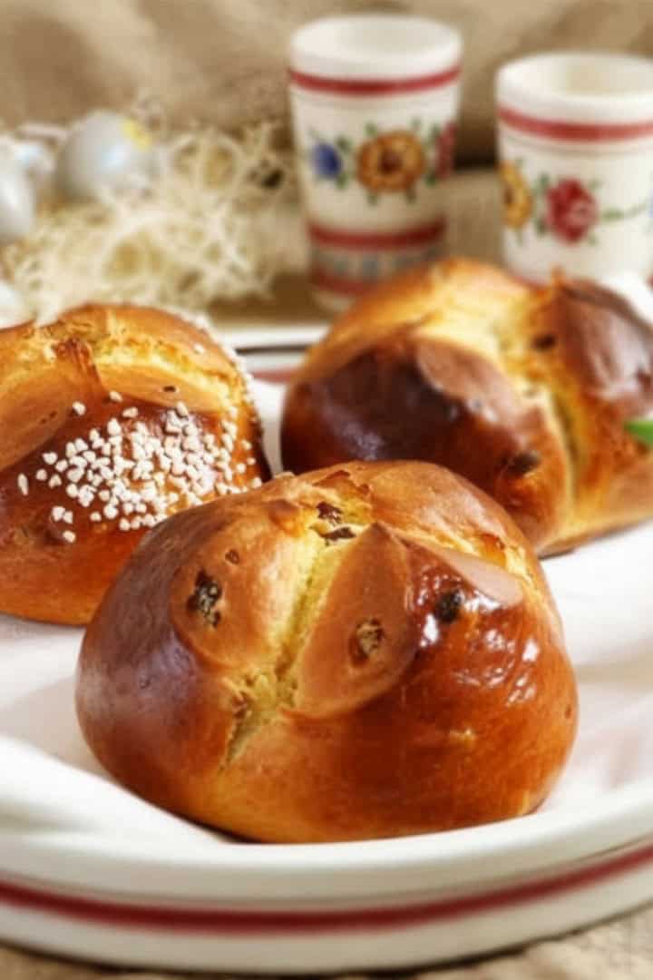 Three golden brown Easter Bread buns with sugar topping rest on a white plate, with decorated cups and a tulip flower in the background.