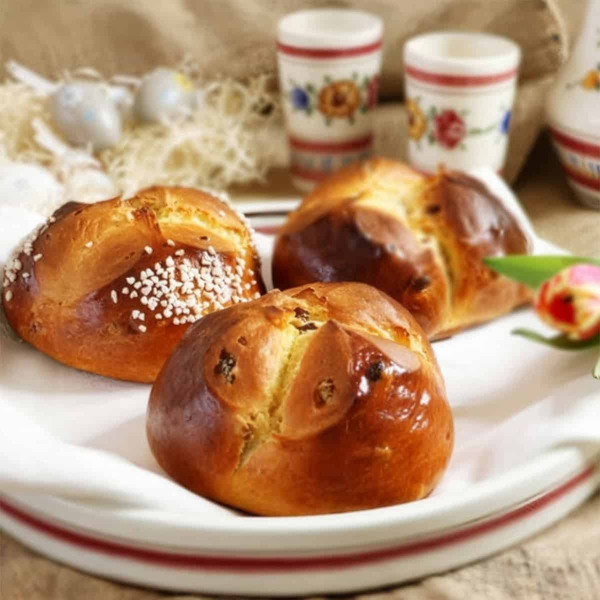 Three golden brown Easter Bread buns with sugar topping rest on a white plate, with decorated cups and a tulip flower in the background.