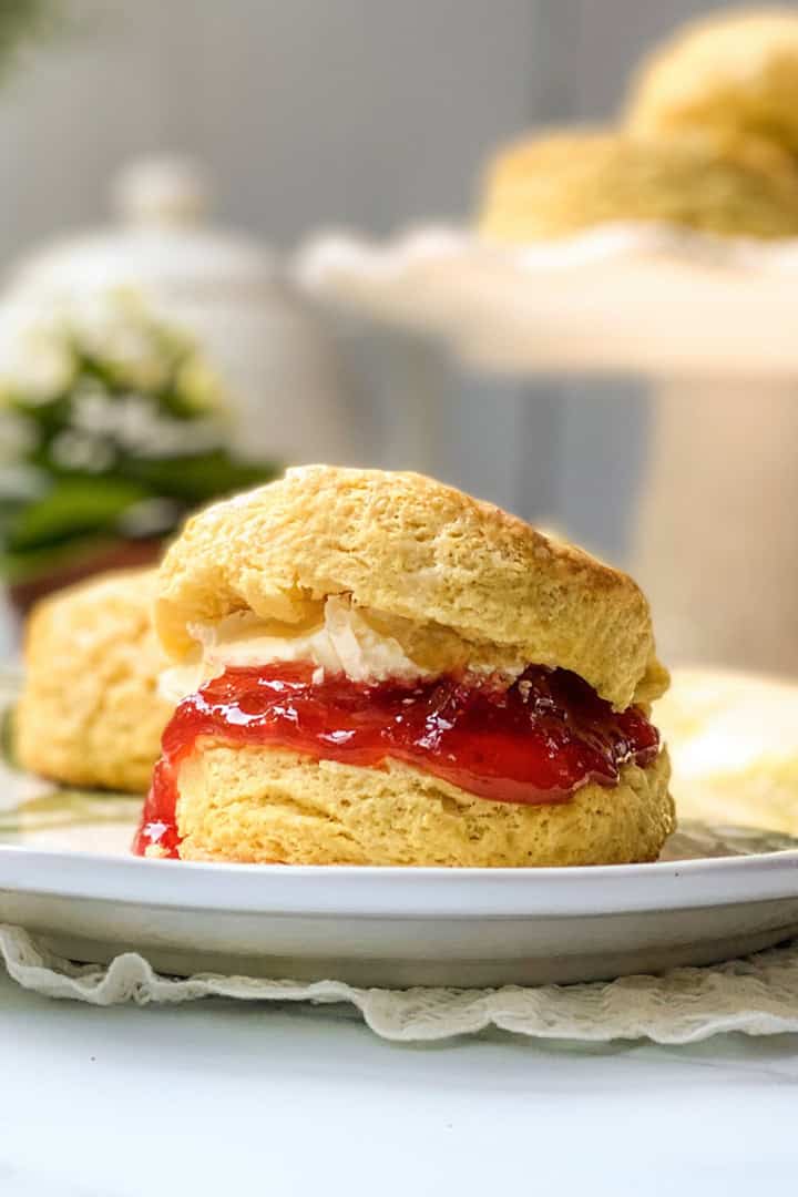 A scone split in half with red jam and cream filling, served on a white plate with another scone and a cake stand in the background-perfectly capturing the charm of a classic Irish scones recipe.