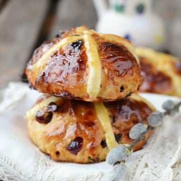 Two glazed hot cross buns with raisins are stacked on a white cloth, highlighting the irresistible results of a perfect hot cross buns recipe, while a branch of pussy willow rests in the foreground.