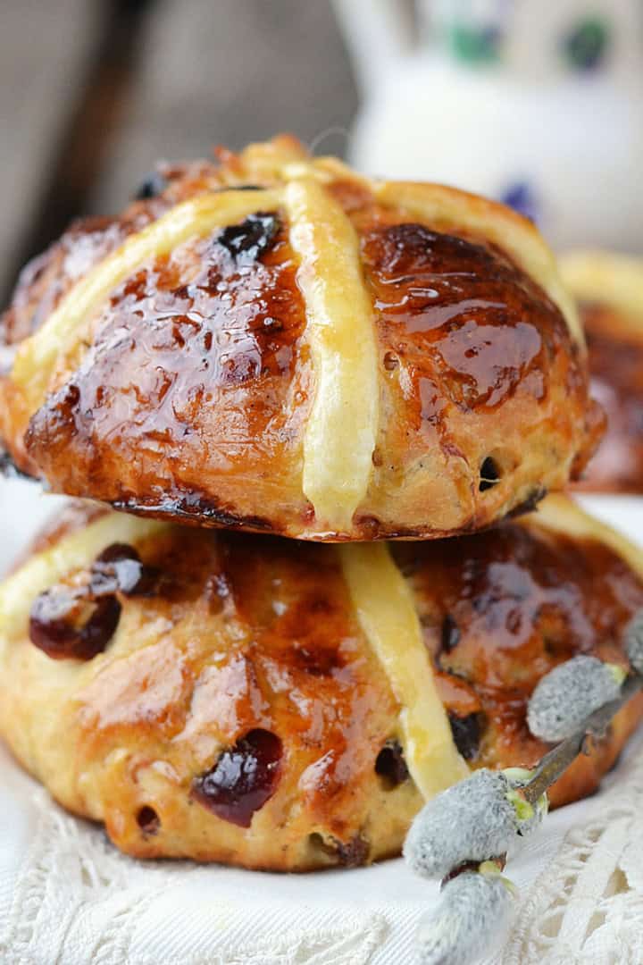 Two glazed hot cross buns with raisins are stacked on a white cloth, highlighting the irresistible results of a perfect hot cross buns recipe, while a branch of pussy willow rests in the foreground.