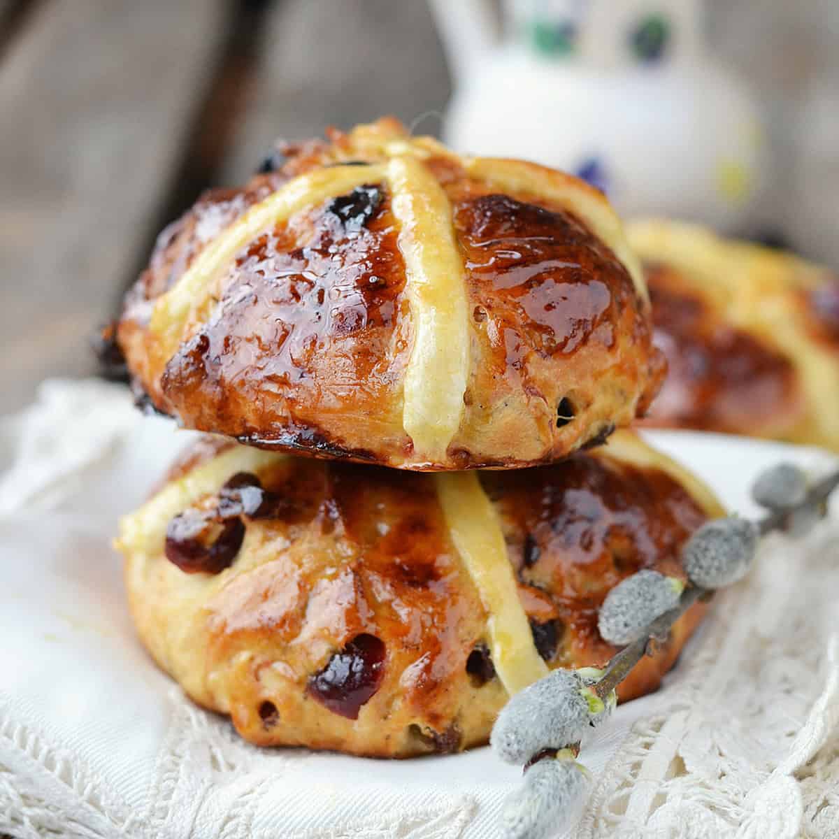 Two glazed hot cross buns with raisins are stacked on a white cloth, highlighting the irresistible results of a perfect hot cross buns recipe, while a branch of pussy willow rests in the foreground.