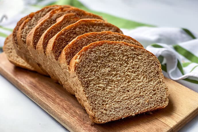 Slices of 100% whole wheat bread are neatly arranged on a wooden cutting board. A green and white striped cloth is partially visible in the background on a light-colored surface.