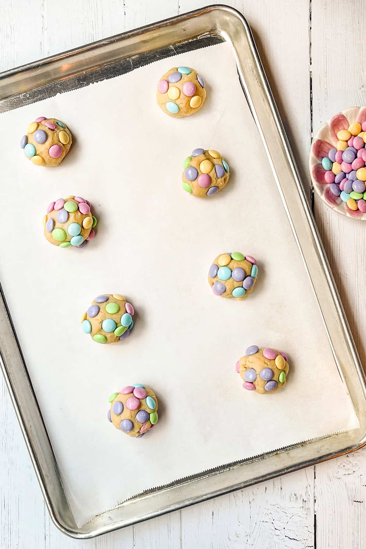 A baking tray lined with parchment paper holds eight unbaked M&M Cookies, each topped with colorful candy-coated chocolates. A bowl of extra candies is visible on the side.
