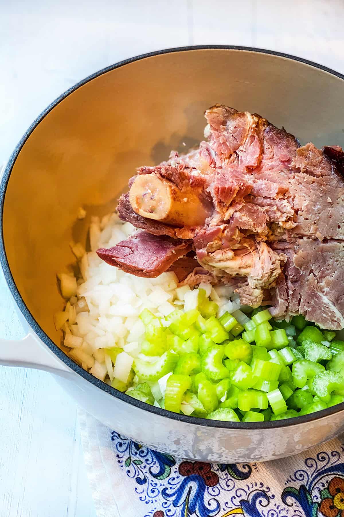 Chopped onions, celery, and a meaty ham bone are placed in a large pot on a patterned cloth, ready to create a hearty ham bone soup.