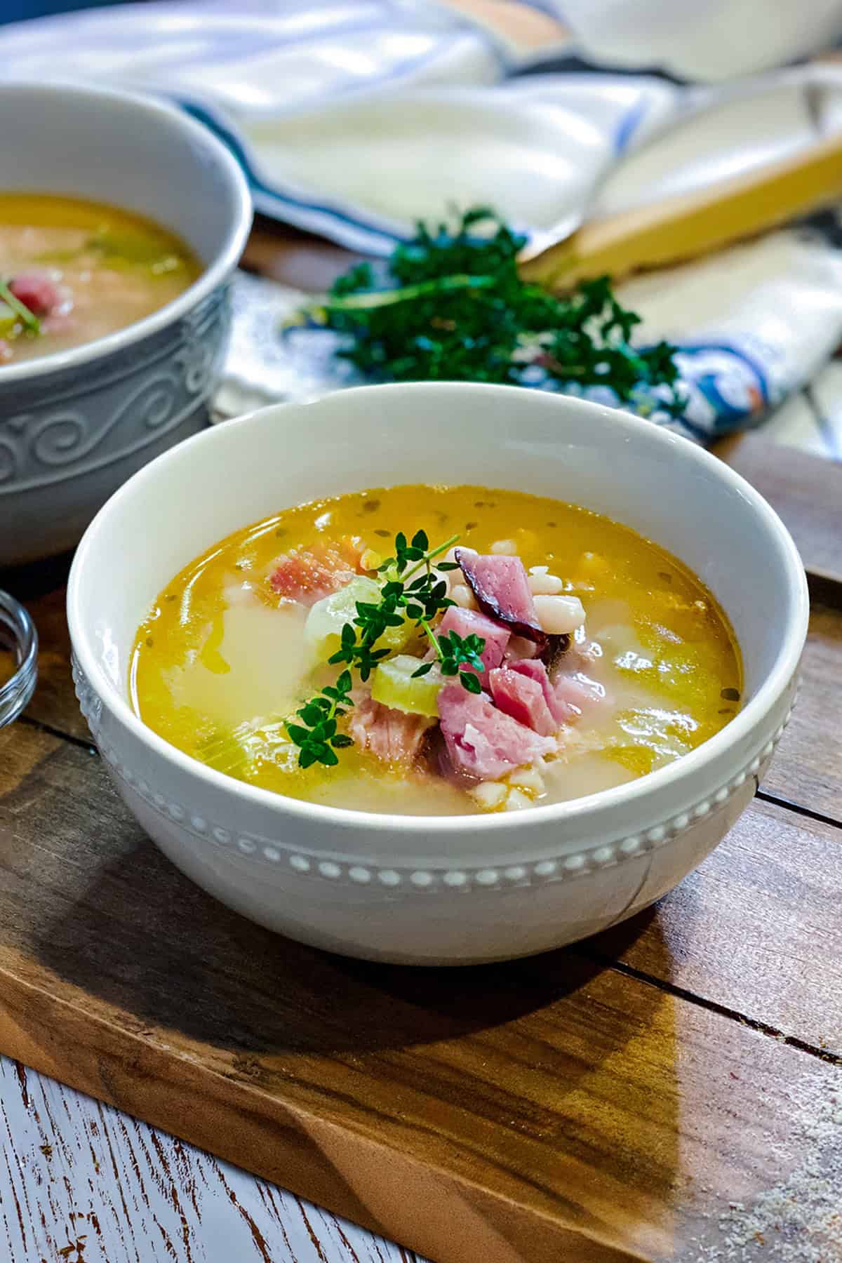 Serving of Ham Bone with white beans in a white bowl topped with fresh thyme leaves rests on a wooden serving tray with more bowls of soup in the background.