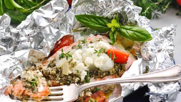 A Spring 5 Ingredient Dinners favorite: a foil packet with baked fish, tomatoes, herbs, and cheese on a blue plate, surrounded by fresh tomatoes, onion, and basil in the background.