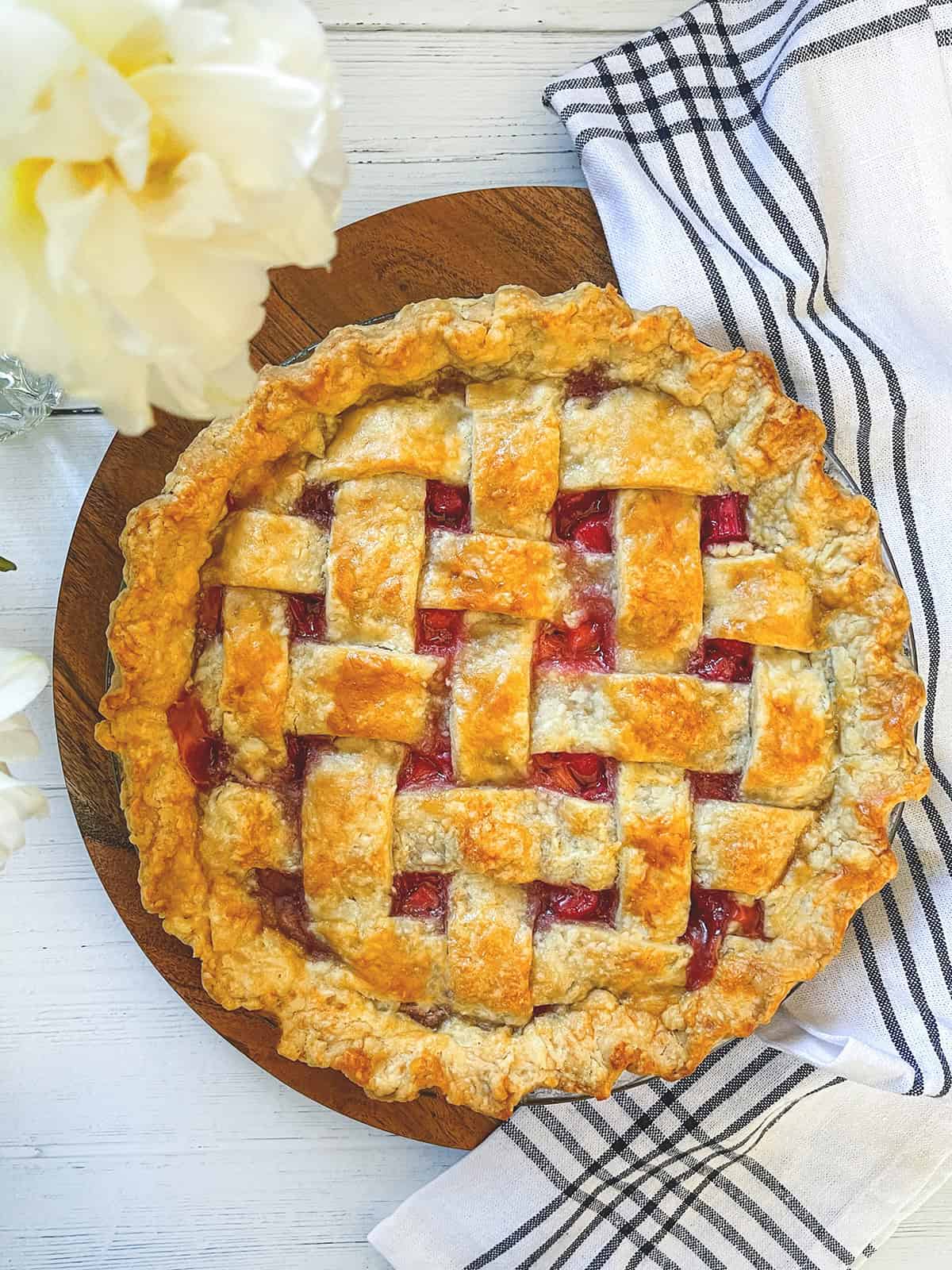 A lattice-top rhubarb pie with a golden crust sits on a wooden board, next to a striped towel and white flowers on a white surface.
