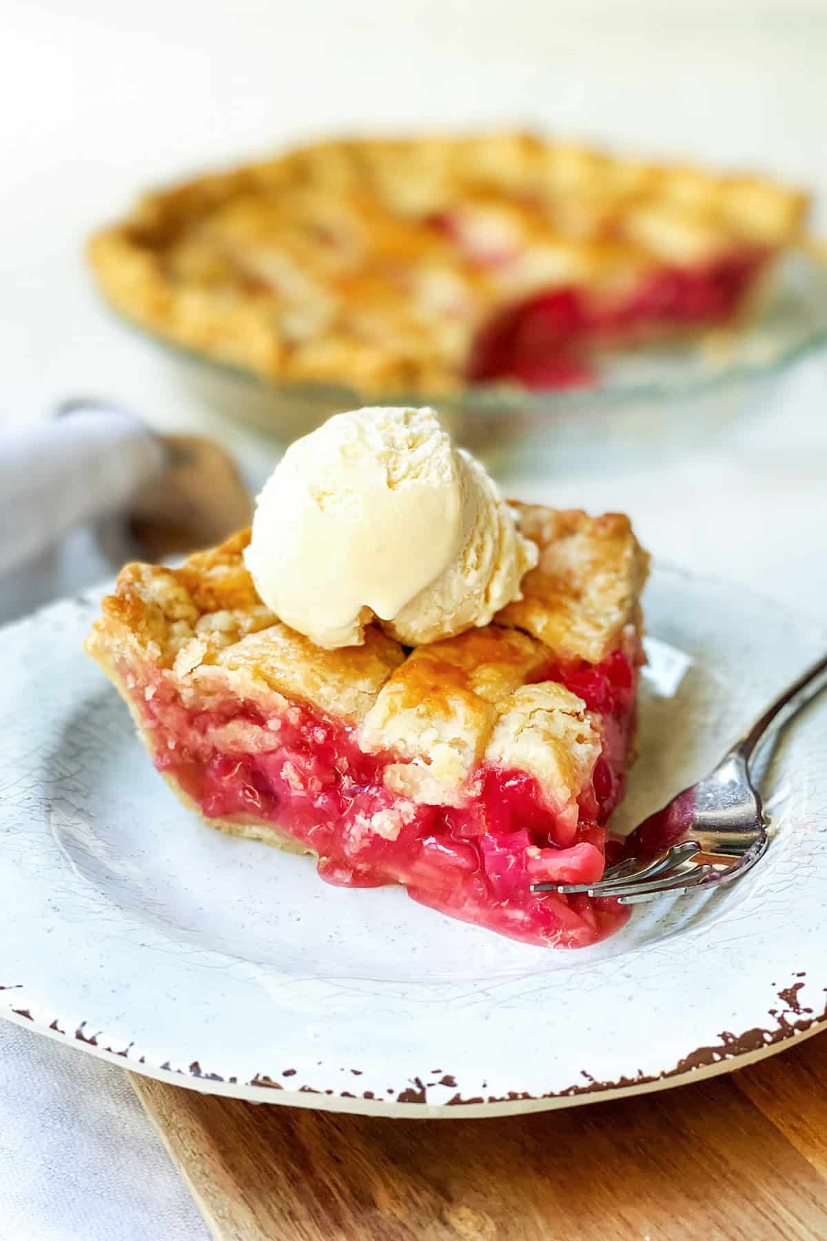 A slice of Rhubarb Pie with a lattice crust is served on a plate, topped with vanilla ice cream and a fork beside it. The rest of the Rhubarb Pie sits temptingly in the background.