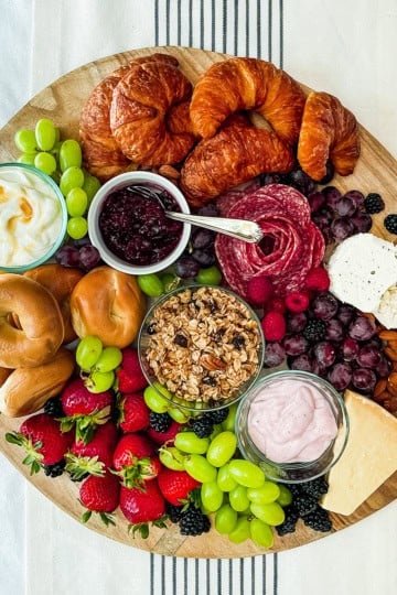 Round charcuterie wooden board filled with brunch foods like croissants, bagels, fruits, meats, and spreads.
