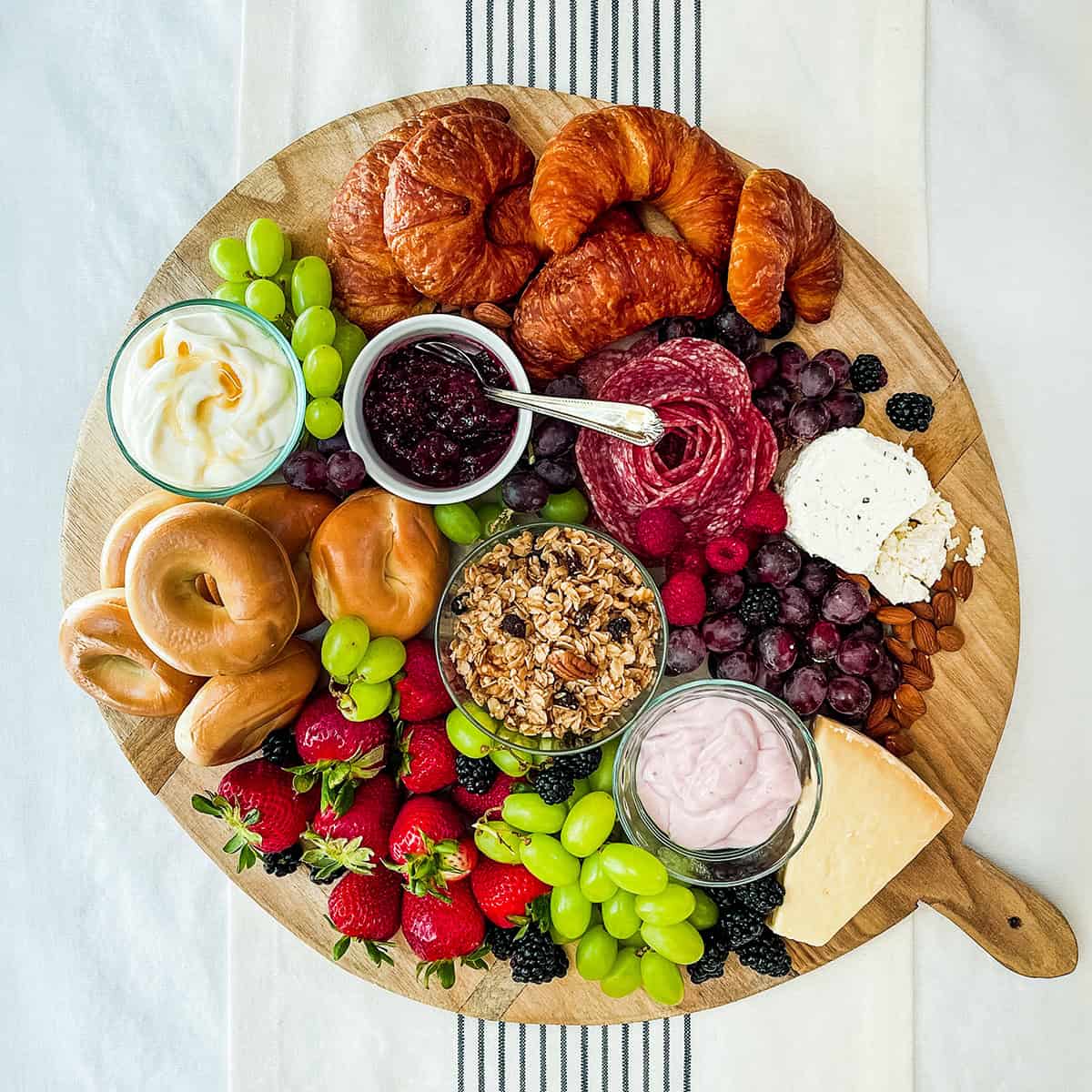 Round charcuterie wooden board filled with brunch foods like croissants, bagels, fruits, meats, and spreads.