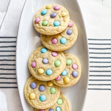 Five Easter M&M Cookies topped with pastel-colored candy-coated chocolates, arranged on a white oval plate.