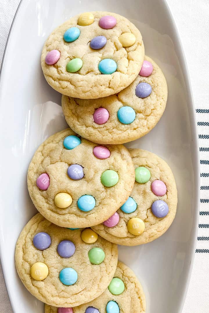 Five Easter M&M Cookies topped with pastel-colored candy-coated chocolates, arranged on a white oval plate.