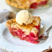 A slice of cherry pie with a lattice crust, reminiscent of classic rhubarb pie, is topped with a scoop of vanilla ice cream and sits on a white plate with a fork.