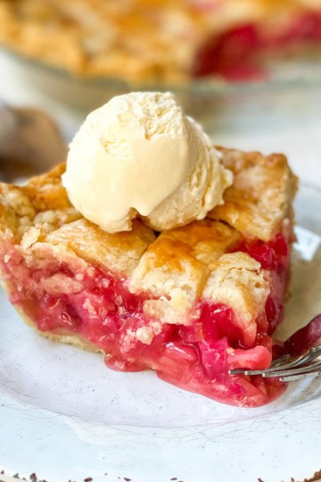 A slice of cherry pie with a lattice crust, reminiscent of classic rhubarb pie, is topped with a scoop of vanilla ice cream and sits on a white plate with a fork.