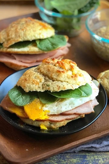 A Scottish Breakfast Sandwich with ham, egg, and spinach on a biscuit sits on a black plate. More sandwiches, a bowl of greens, and mustard are in the background.