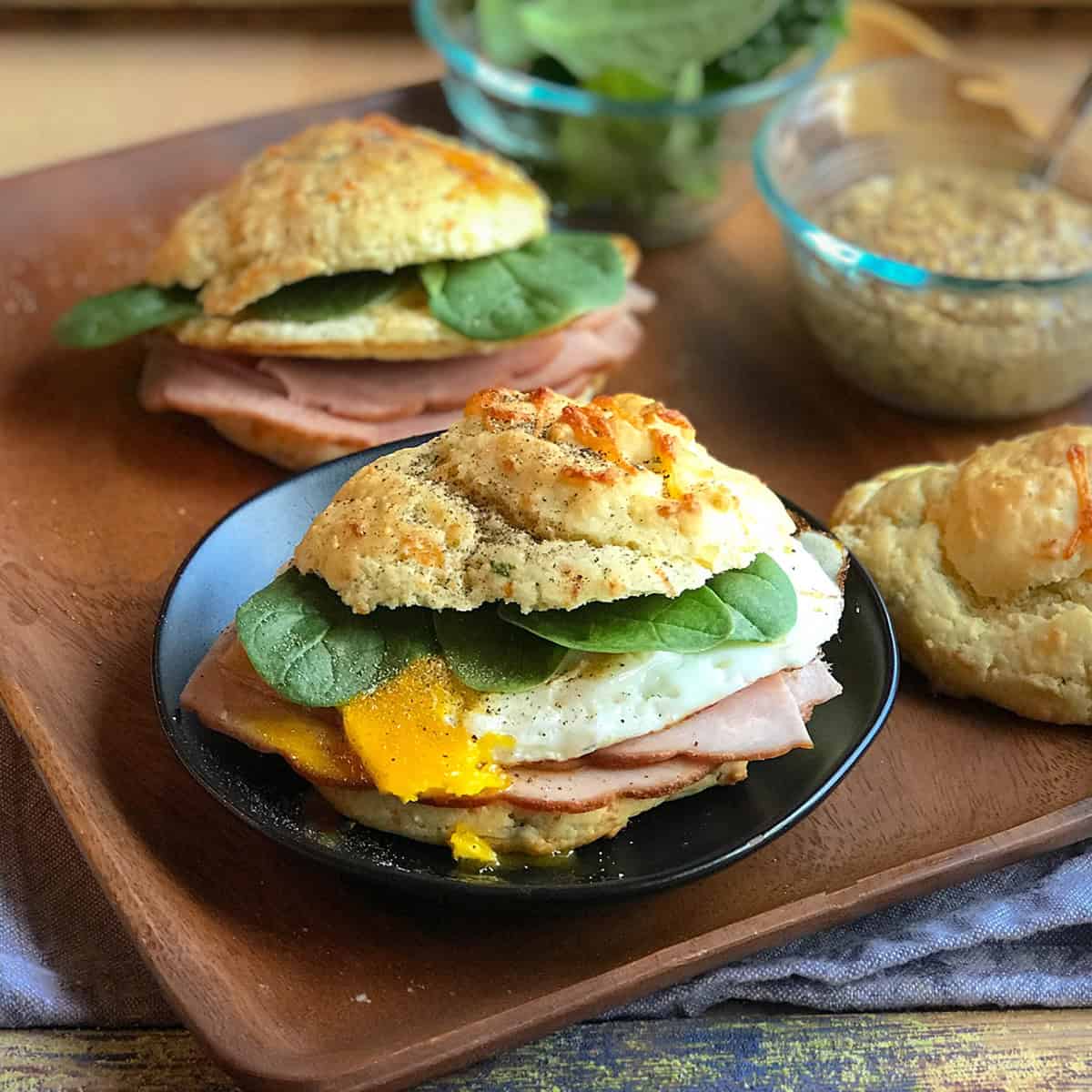 A Scottish Breakfast Sandwich with ham, egg, and spinach on a biscuit sits on a black plate. More sandwiches, a bowl of greens, and mustard are in the background.