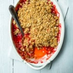 A white baking dish with strawberry crisp, partially served with a spoon, reveals a layer of vibrant red fruit filling beneath a golden oat topping.