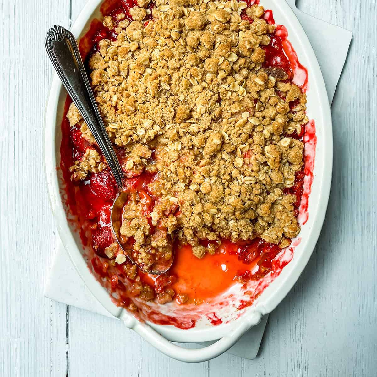 A white baking dish with strawberry crisp, partially served with a spoon, reveals a layer of vibrant red fruit filling beneath a golden oat topping.