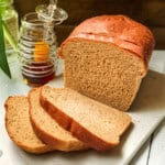 A loaf of whole wheat bread with three slices cut, placed on a white cutting board next to a jar of honey with a honey dipper inside.