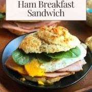 A Scottish Breakfast Sandwich featuring ham, a fried egg, spinach, and a cheddar biscuit is served on a black plate, labeled as Scottish inspired Cheddar Biscuit & Ham Breakfast Sandwich.